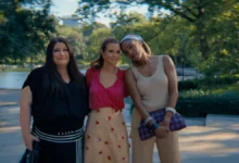 "Three women standing close together outdoors on a sunny day in a lush park, smiling and posing for a photo with trees, greenery, and a body of water in the blurred background."