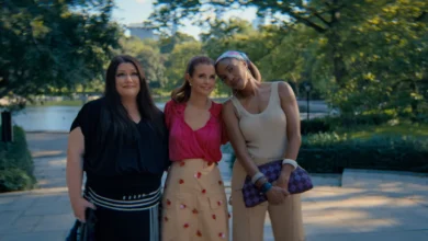 "Three women standing close together outdoors on a sunny day in a lush park, smiling and posing for a photo with trees, greenery, and a body of water in the blurred background."