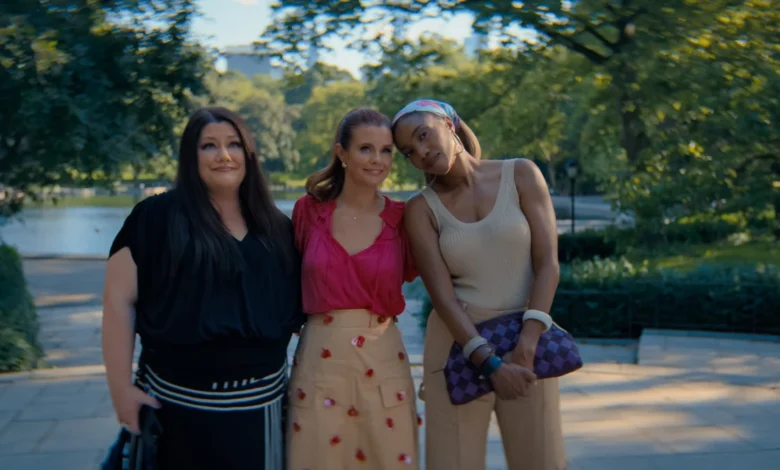 "Three women standing close together outdoors on a sunny day in a lush park, smiling and posing for a photo with trees, greenery, and a body of water in the blurred background."