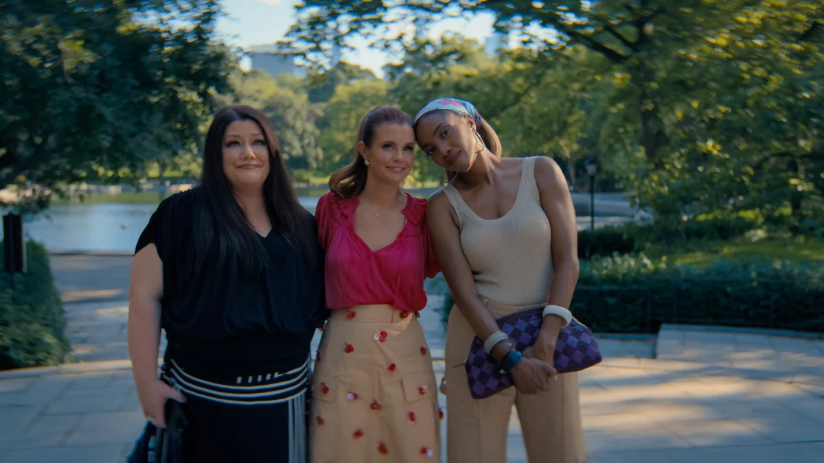 "Three women standing close together outdoors on a sunny day in a lush park, smiling and posing for a photo with trees, greenery, and a body of water in the blurred background."